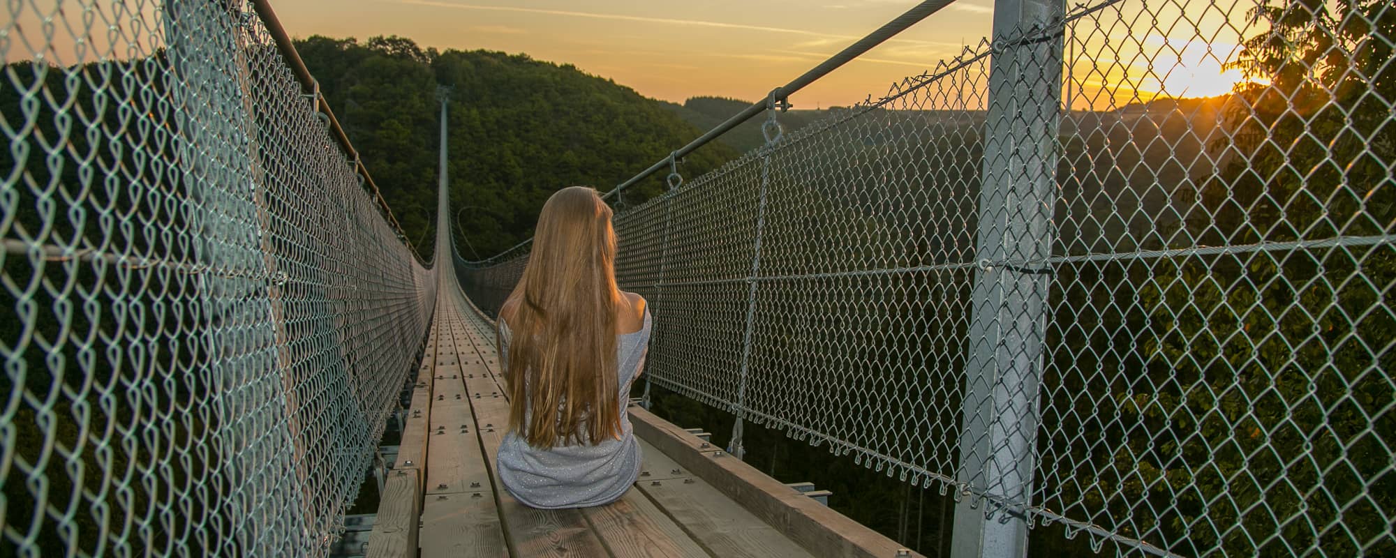 Frau mit langen Haaren sitz bei Sonnenaufgang auf der Geierlay Hängeseilbrücke