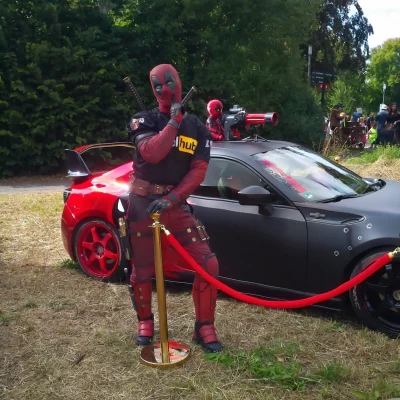 Person in Deadpool-Kostüm lehnt an Absperrpfosten mit rotem Seil vor einem schwarzen Sportwagen mit roten Felgen und einem weiteren Deadpool-Figur auf dem Autodach