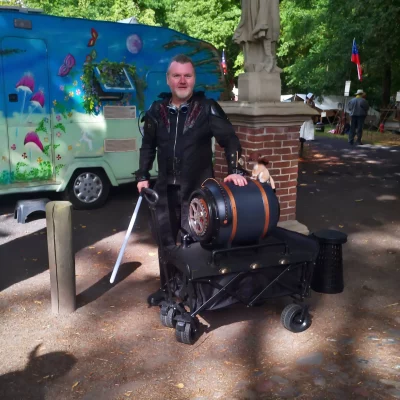 Mann mit schwarzer Jacke steht neben einem fahrbaren Gerät mit einem Fass und einem kleinen Tier darauf in einem Park mit bemaltem Wohnwagen im Hintergrund