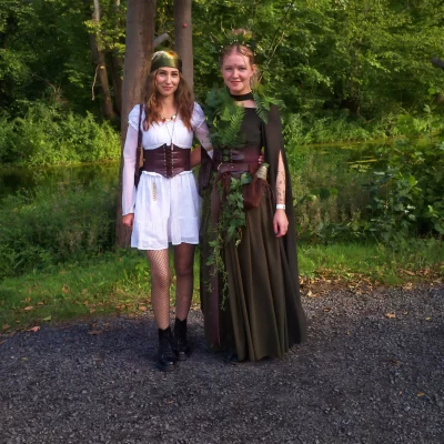 Zwei Frauen in mittelalterlich inspirierten Kostümen stehen nebeneinander vor einem Baum in einem Waldgebiet
