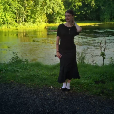 Frau in schwarzem Kleid und weißen Socken steht am Ufer eines mit Pflanzen bewachsenen Teichs und berührt ihr Haar