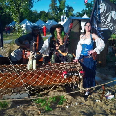 Drei Personen in Piratenkostümen vor einem hölzernen Stand mit Netz und Kerzen, im Hintergrund weiße Zelte und eine schwarze Flagge mit Einhornmotiv