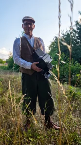 Fotograf Alexander Konopka mit Schiebermütze, weißem Hemd und brauner Weste steht auf einer Wiese und hält seine Graflex Kamera