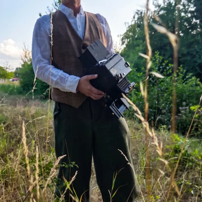 Fotograf Alexander Konopka mit Schiebermütze, weißem Hemd und brauner Weste steht auf einer Wiese und hält seine Graflex Kamera