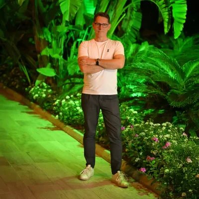 Alexander mit Brille, weißem T-Shirt und dunkler Hose steht mit verschränkten Armen auf einem Weg neben einer bepflanzten Fläche mit grünen Blättern und Blumen