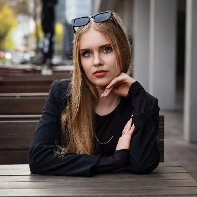 Nahaufnahme einer jungen Frau mit langem blonden Haar und schwarzem Blazer an einem Holztisch. Ausdrucksstarker Blick, eckige Sonnenbrille im Haar und sanftes Bokeh im urbanen Hintergrund.