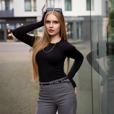 Frau mit langen, glatten Haaren und Sonnenbrille auf dem Kopf steht mit einer Hand am Kopf und trägt ein schwarzes Oberteil sowie graue Hose vor einer Glaswand im Medienhafen Düsseldorf.