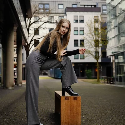 Weibliches Model mit langen Haaren in schwarzem Oberteil und grauer Hose steht mit einem Bein auf einer Holzbank in einem urbanen Innenhof