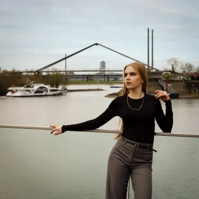 Frau mit langem Haar in schwarzem Oberteil und grauer Hose lehnt an einer Brüstung vor dem Rhein mit einer markanten Brücke und einem Boot im Hintergrund