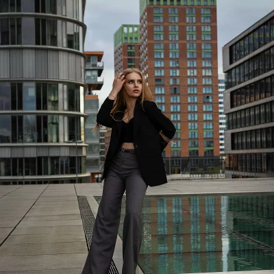 Model Anastasiia mit der rechten Hand in ihren langen Haaren und schwarzem Blazer steht auf einem modernen Platz vor hohen Gebäuden und einem Wasserbecken bei einem TFP Fotoshooting in Düsseldorf