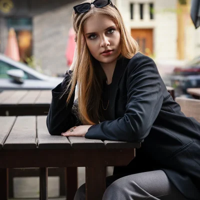 Urbanes Portrait von Anastasiia in schwarzem Blazer und grauer Hose, die Unterarme auf einen Holztisch in einem Straßencafé gelehnt, Sonnenbrille im Haar, direkter Blick in die Kamera, im Hintergrund Häuserfassade und parkende Autos