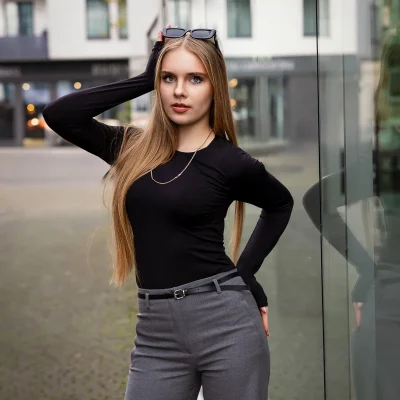 Frau mit langen, glatten Haaren und Sonnenbrille auf dem Kopf steht mit einer Hand am Kopf und trägt ein schwarzes Oberteil sowie graue Hose vor einer Glaswand im Medienhafen Düsseldorf.