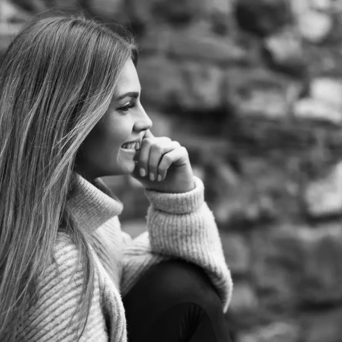 Portrait eines lächelnden weiblichen Models, das seitlich sitzt, den Ellenbogen auf dem Knie stützt, vor einer unscharfen Mauer