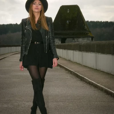 Frau in schwarzem Kleid, Lederjacke, Hut und Overknee-Stiefeln unterwegs auf der Staumauer am Möhnesee bei bewölktem Himmel