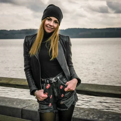 Frau in schwarzer Beanie, Lederjacke und kurzer Jeans mit Rolling-Stones-Zungen-Logos, stehend am See