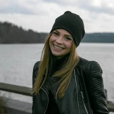 Anna Katharina Klein mit schwarzer Mütze und Lederjacke lächelt vor einem See im Hintergrund.