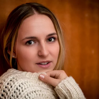 Porträt einer Frau mit braunen Augen, die über die Schulter schaut und einen weißen, grob gestrickten Pulli trägt.