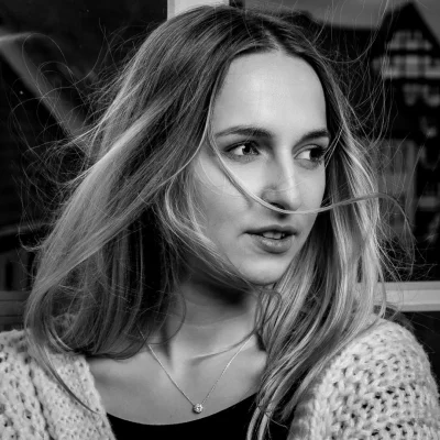 Frau mit locker fallenden Haaren, die seitlich blickt, trägt ein grob gestricktes Oberteil vor einem Fenster in Schwarz-Weiß.