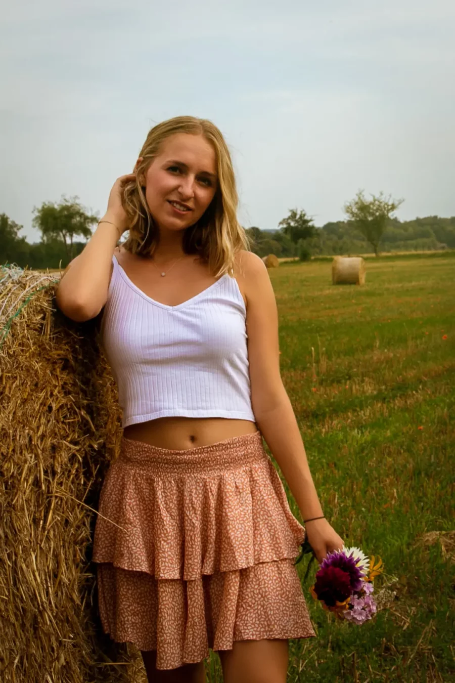Frau mit lockerem, weißen Top und gemustertem, zweilagigem Rock lehnt an rundem Heuballen und hält einen kleinen Blumenstrauß.