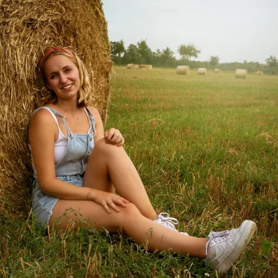 Frau in kurzer Jeanslatzhose und mit rotem Haarband sitzt lächelnd und entspannt am Fuß eines großen Heuballens auf einem Feld.