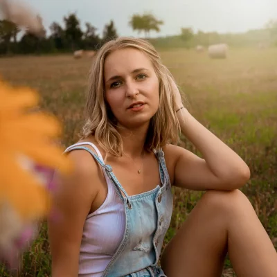 Anna Sommer sitzt auf einem Feld, trägt ein weißes Top mit blauer Latzhose und blickt mit entspannter Haltung in die Kamera.