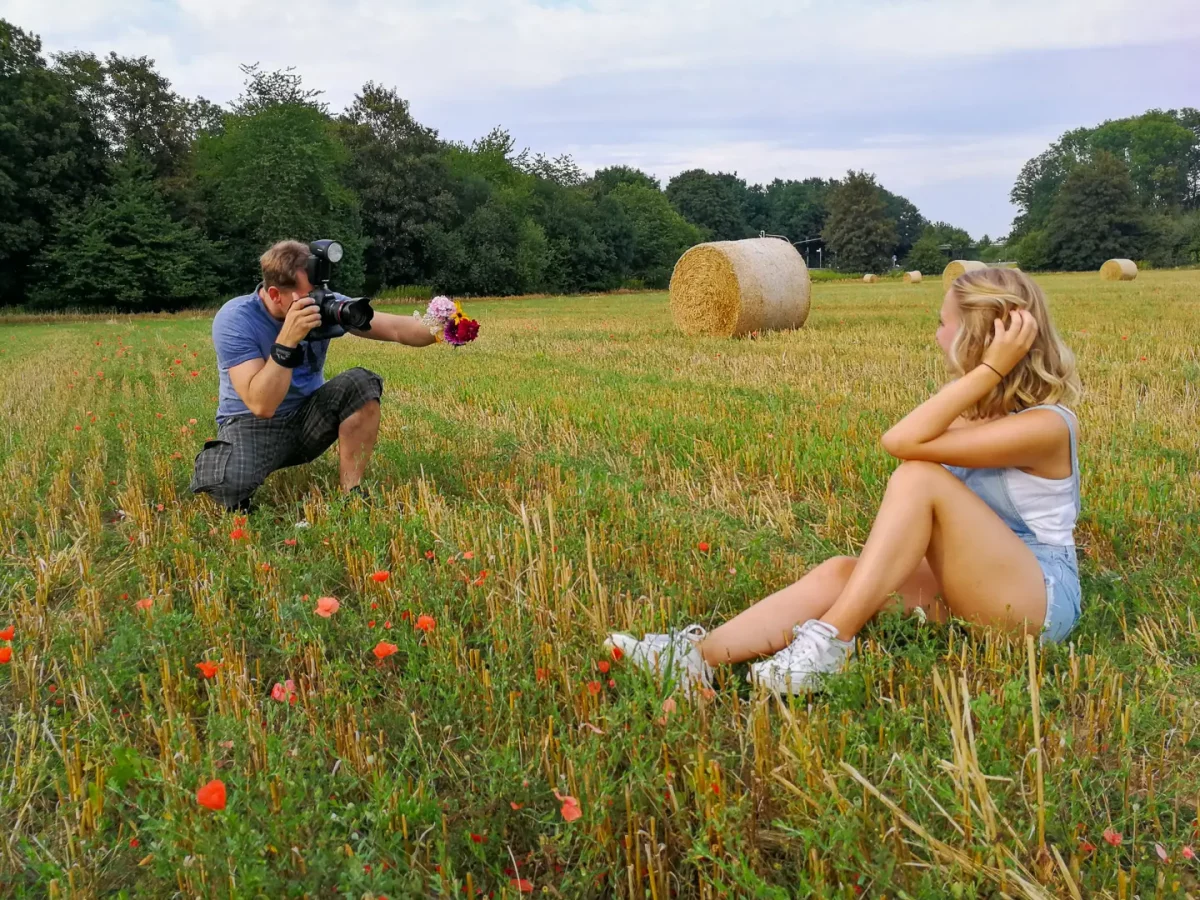 Fotograf hält Blumenstrauß und fotografiert ein sitzendes weibliches Model in Shorts und weißem Top auf Feld mit Strohballen.