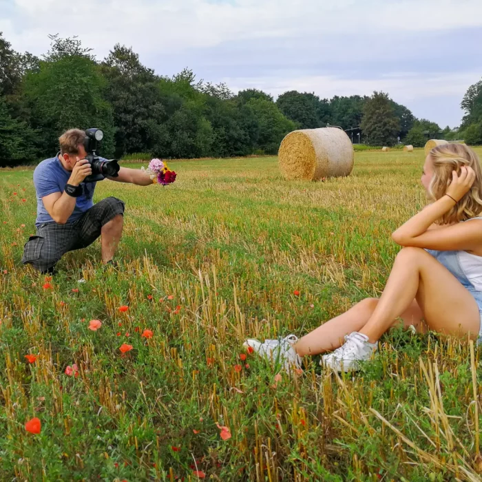 TFP Fotoshooting Making-Of: Fotograf Alexander Konopka hält Blumenstrauß und fotografiert ein sitzendes weibliches Model in Shorts und weißem Top auf Feld mit Strohballen.
