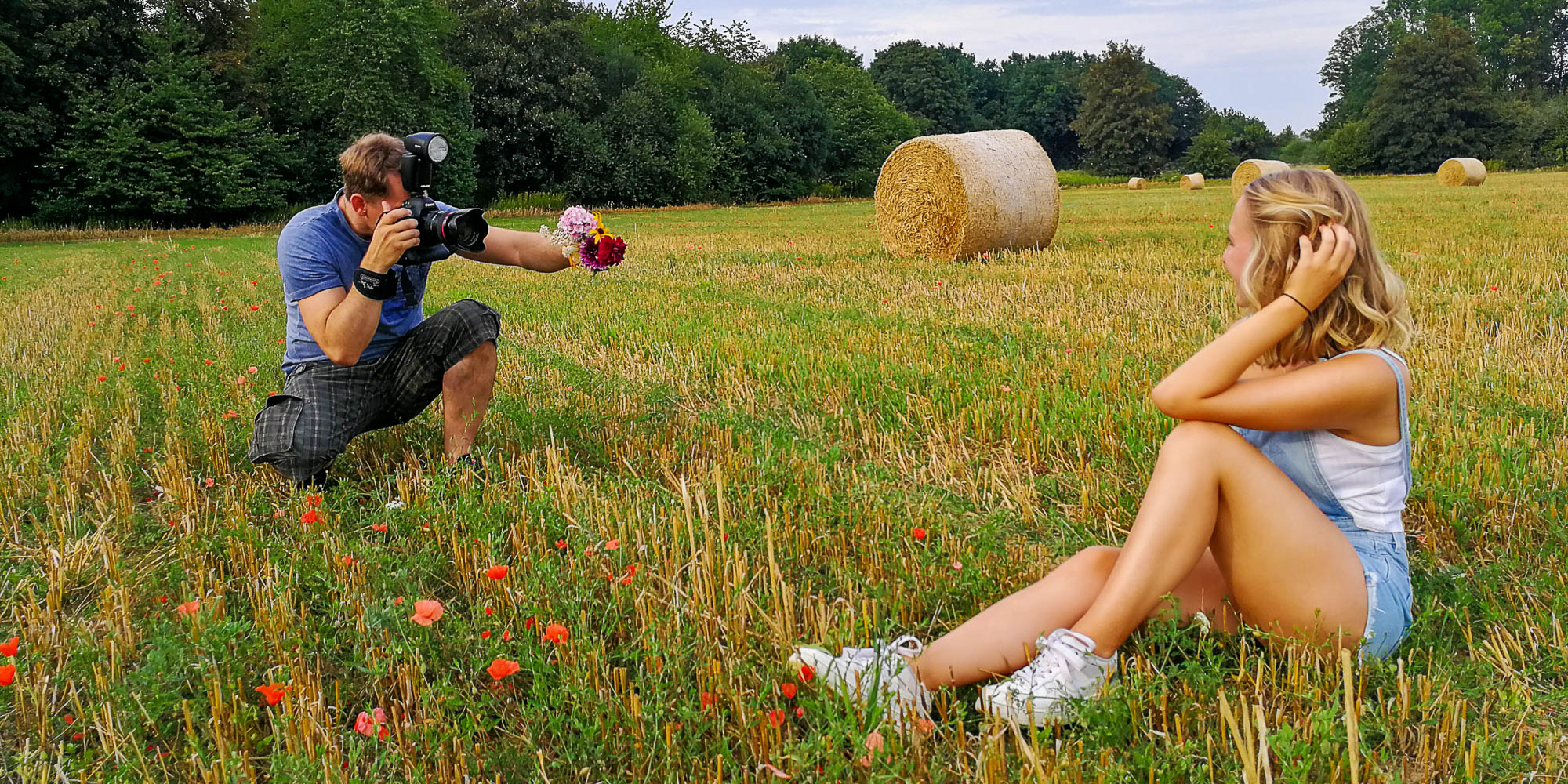 TFP Fotoshooting Making-Of: Fotograf Alexander Konopka hält Blumenstrauß und fotografiert ein sitzendes weibliches Model in Shorts und weißem Top auf Feld mit Strohballen.