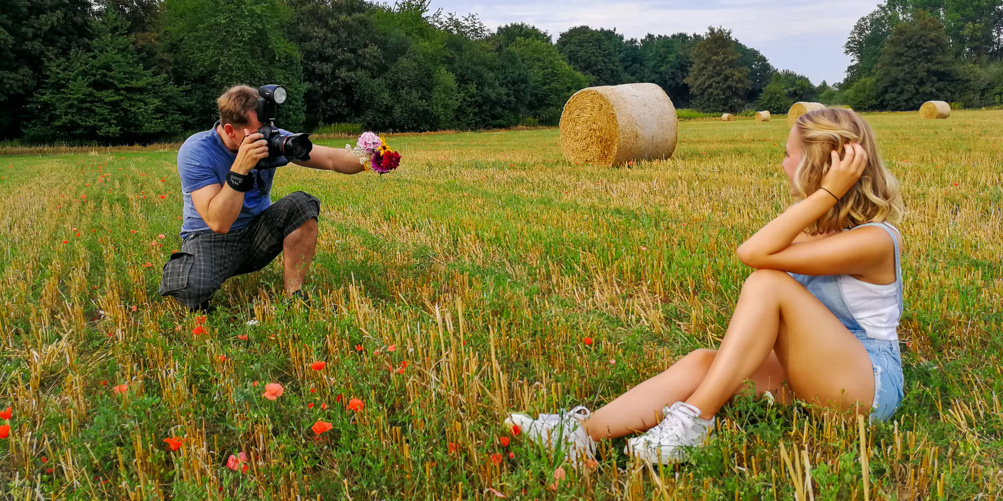 TFP Fotoshooting Making-Of: Fotograf Alexander Konopka hält Blumenstrauß und fotografiert ein sitzendes weibliches Model in Shorts und weißem Top auf Feld mit Strohballen.