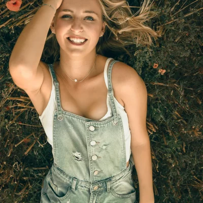 Frau in Jeanslatzhose und einem weißen Trägershirt liegt lächelnd auf dem Rücken im Gras, eine Hand berührt das blonde Haar.