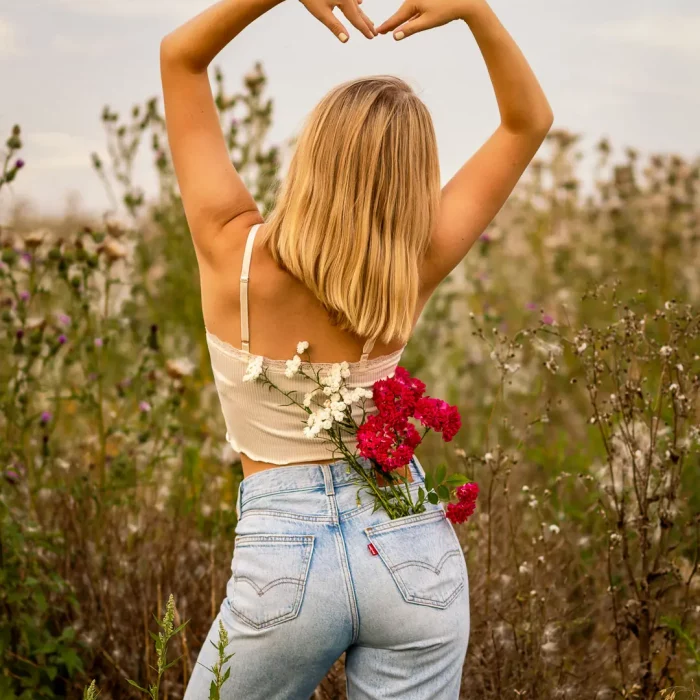 Blonde Frau formt mit erhobenen Armen ein Herz, trägt weißes Top und blaue Jeans mit Blumen in der Gesäßtasche, vor Wildkräuterfeld