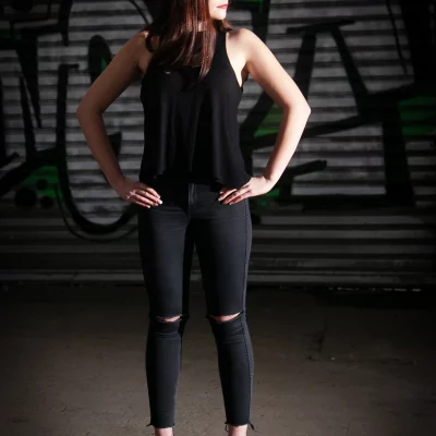 Annika steht mit Händen in der Hüfte vor einer mit Graffiti besprühten Metalljalousie und trägt schwarzes ärmelloses Oberteil, schwarze enge Jeans mit Löchern an den Knien und schwarze High Heels