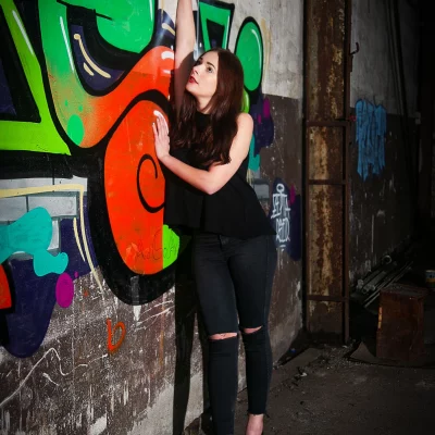 Frau in schwarzer Kleidung und High Heels lehnt an einer Wand mit buntem Graffiti in einem verlassenen Industriegebäude