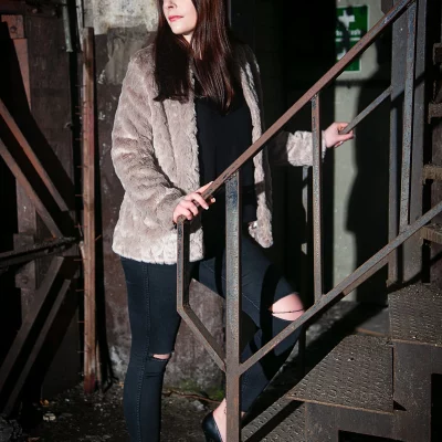 Frau mit langem Haar, schwarzer Hose mit Rissen und hellgrauem Fellmantel steht auf einer rostigen Metalltreppe in einem verlassenen Gebäude