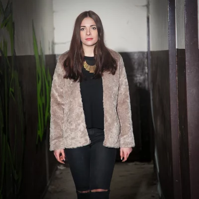 Frau mit langem braunem Haar trägt eine helle Felljacke, schwarze Hose mit Rissen an den Knien und eine auffällige goldfarbene Halskette in einem dunklen, schmalen Gang.