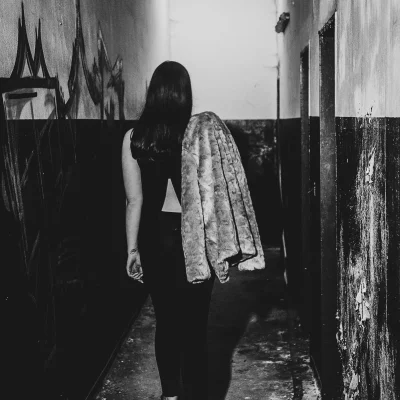 Frau mit langen Haaren und hochhackigen Schuhen geht in einem schmalen, abgenutzten Gang, trägt eine Jacke über dem Arm; Lost Place im Sauerland