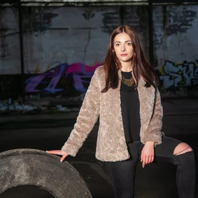 Frau mit langem Haar, beiger Felljacke und schwarzer Kleidung lehnt mit einer Hand auf einem großen Reifen in einer verlassenen Halle mit Graffiti an der Wand