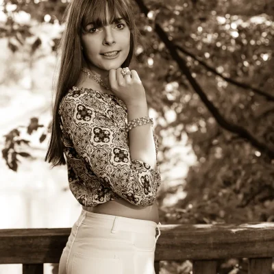 Model Annika mit langen Haaren und gemustertem Oberteil steht seitlich an einem Holzzaun vor unscharfem Laubwerk