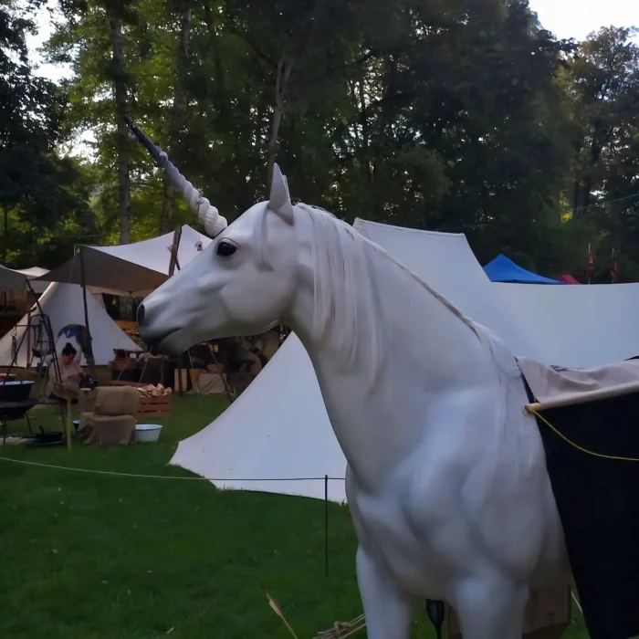 Lebensgroße weiße Einhornfigur mit langem Horn vor mehreren Zelten in einem bewaldeten Freiluftlager