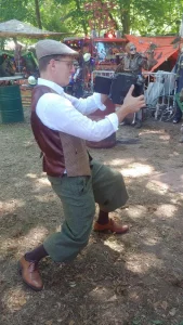 Fotograf in Vintage-Kleidung mit Schiebermütze, Knickerbocker-Hose und Weste hält eine Graflex-Kamera auf einem Fantasy-Festival