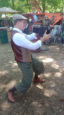 Fotograf in Vintage-Kleidung mit Schiebermütze, Knickerbocker-Hose und Weste hält eine Graflex-Kamera auf einem Fantasy-Festival