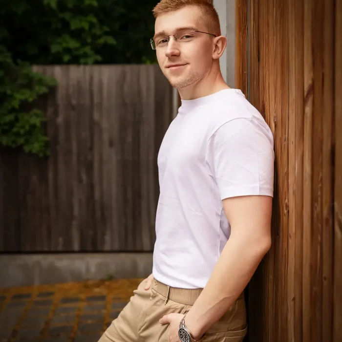 Junges männliches Model mit Brille lehnt entspannt an Holzwand, trägt weißes T-Shirt, beige Hose und Armbanduhr.