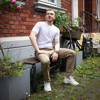 Mann mit Brille, lässig sitzend auf einer Holzbank vor einem Backsteinhaus, trägt weißes T-Shirt, beige Hose und weiße Sneaker.
