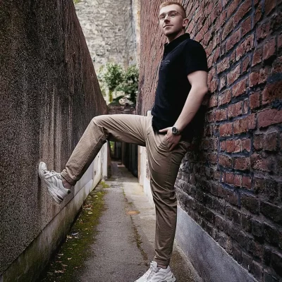 Mann lehnt mit einer Hand in der Hosentasche und einem Bein an einer Mauer in enger Gasse, trägt schwarzes Poloshirt und beige Hose.