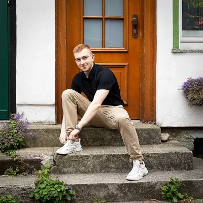 Junger Mann sitzt auf einer Außentreppe vor Holztür und schnürt weiße Sportschuhe mit beigefarbener Hose und schwarzem Poloshirt.