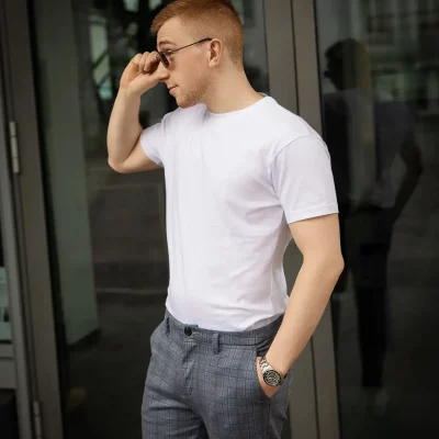 Ansgar Schwulst steht mit Sonnenbrille vor einer Glastür und trägt ein weißes T-Shirt sowie eine grau karierte Hose.