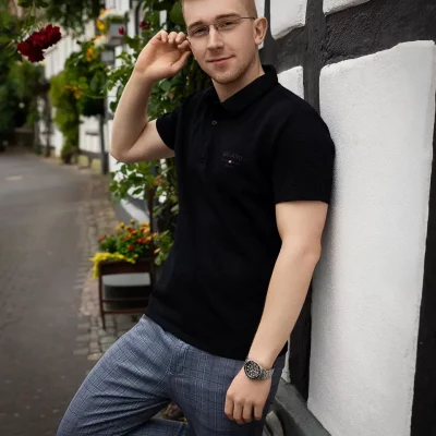 Mann lehnt lässig an einem Fachwerkhaus in Soest, trägt schwarzes Poloshirt und karierte Hose, hinter ihm Blumen und grünes Laub.