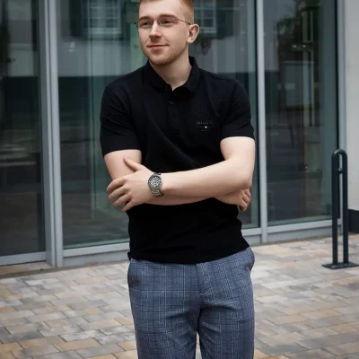 Mann mit Brille, schwarzem Poloshirt und karierten Hosen steht mit verschränkten Armen vor einem modernen Gebäude in Soest.
