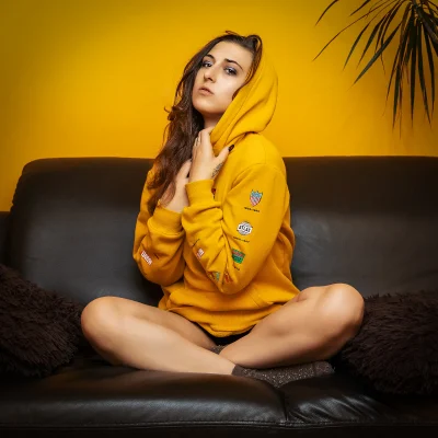 Weibliches Model sitzt mit verschränkten Beinen auf schwarzem Ledersofa, trägt gelben Kapuzenpullover mit Aufnähern.