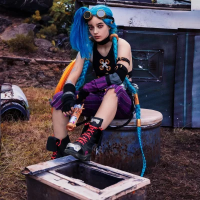 Cosplay-Fotograf NRW: Jinx mit blauen Zöpfen in Punk-Kleidung sitzt auf einem rostigen Fass und hält eine bunte Spielzeugpistole.